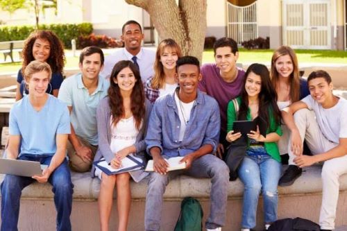 A group of secondary students sits on a concrete bench outside a school on a sunny day.